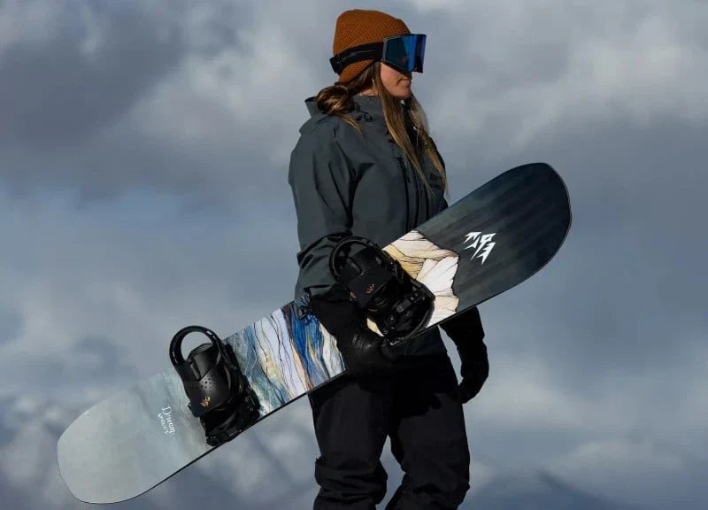 A woman snowboarder holds her board as she wears a hat and goggles.