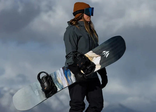 A woman snowboarder holds her board as she wears a hat and goggles.